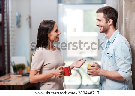 Spending a nice coffee break. Two cheerful young people holding coffee cups and talking while standing in office