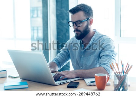 Concentrated on work. Concentrated young beard man working on laptop while sitting at his working place in office