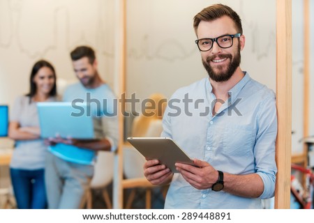 Confident IT expert. Cheerful young man holding digital tablet and smiling while his colleagues working in the background