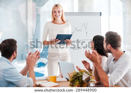Great presentation! Cheerful young woman standing near whiteboard and smiling while her colleagues sitting at the desk and applauding