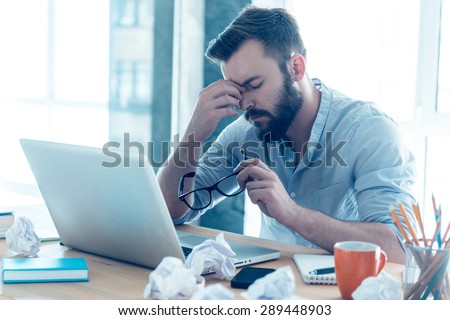Feeling exhausted. Frustrated young beard man massaging his nose and keeping eyes closed while sitting at his working place in office