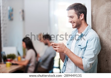 Staying in touch. Happy young man looking at his mobile phone and smiling while his colleagues working in the background
