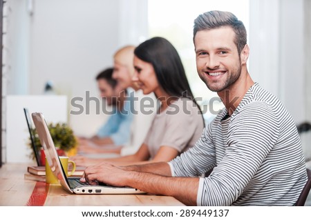 I love my job! Group of cheerful business people in smart casual wear working at their laptops while handsome man looking at camera and smiling