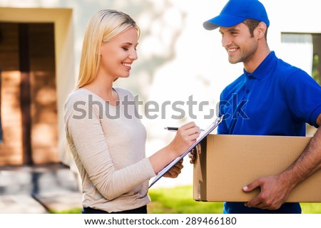 Sign here please! Smiling young delivery man holding a cardboard box while beautiful young woman putting signature in clipboard