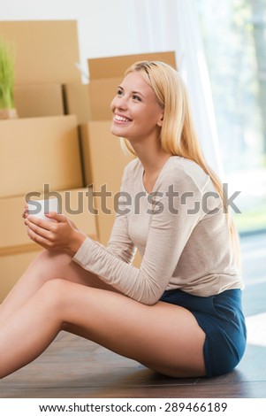 Time for inspiration. Beautiful young woman sitting on the floor and holding cup of coffee while cardboard boxes laying in the background