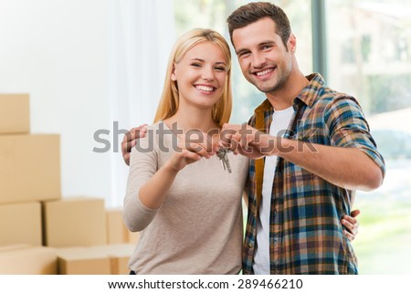 Keys of their new house. Cheerful young couple holding keys and smiling while standing in their new house