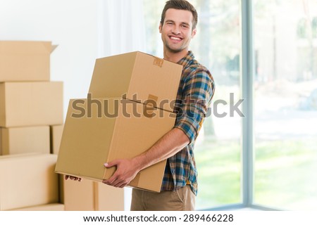 Moving to a new apartment. Cheerful young man holding a cardboard boxes and smiling at camera while other carton boxes laying on background