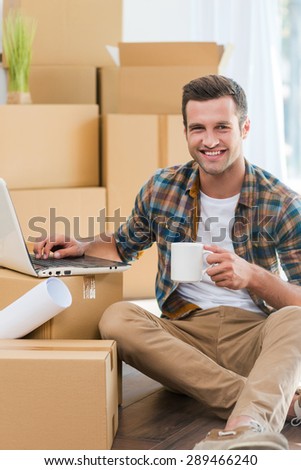 Coffee break. Happy young man sitting on the floor and working on laptop while cardboard boxes laying in the background