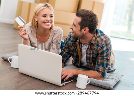Online shopping makes life easier. Smiling young couple laying on the floor of their new apartment and shopping through Internet while cardboard boxes laying in the background