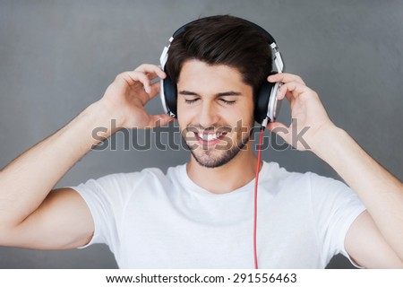Enjoying his favorite music. Handsome young man keeping eyes closed and adjusting his headphones while standing against grey background