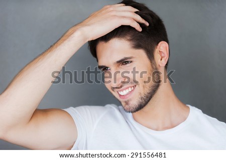 Carefree handsome. Happy young man holding hand in hair and looking away while standing against grey background