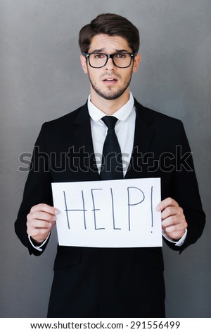 Asking for help. Frustrated young businessman holding paper with text and looking at camera while standing against grey background