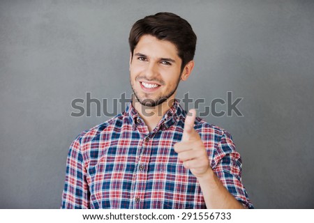 You can do it! Cheerful young man pointing at camera and smiling while standing against grey background