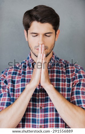 Hoping for good. Thoughtful young man holding hands clasped near face and keeping eyes closed while standing against grey background