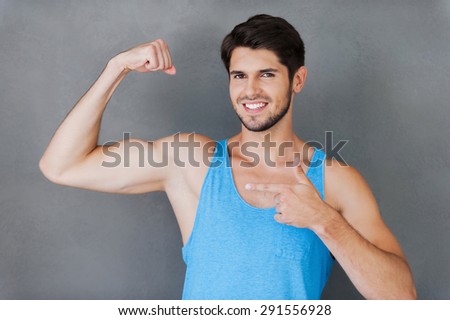 Proud of his perfect biceps. Cheerful young muscular man pointing his bicep while standing against grey background
