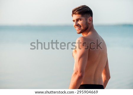 Strong and handsome. Rear view of young muscular man looking over shoulder and smiling while standing at the seaside