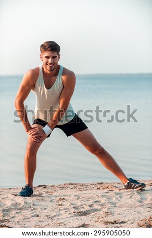 Warming up his muscles. Smiling young muscular man stretching his legs before running while standing outdoors