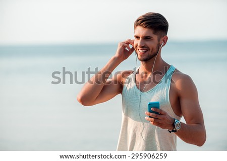 Enjoying music and fresh air. Happy young muscular man in headphones holding smart phone and smiling while standing outdoors