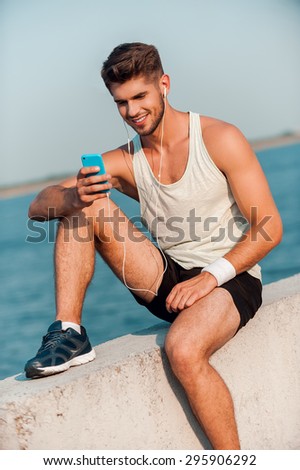 Choosing right music for training. Happy young muscular man in headphones holding smart phone and smiling while sitting on parapet