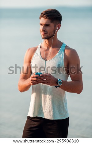Music and beach all I need for running. Confident young muscular man in headphones holding smart phone and looking away while standing outdoors