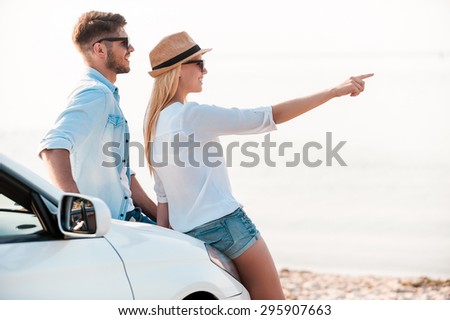 Enjoying nice scenery. Side view of happy young woman pointing away while her boyfriend leaning at the hood of their white convertible