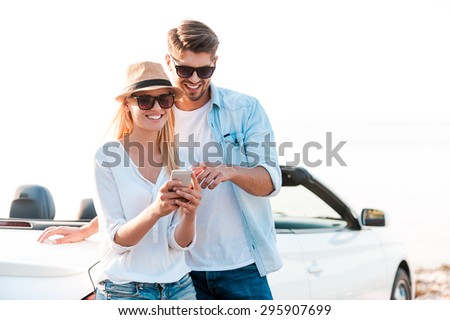 Watching photos of their road trip. Happy young couple looking at mobile phone while leaning at their white convertible