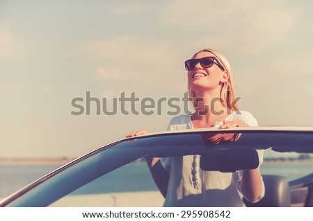 Feeling freedom. Beautiful young woman in eyewear enjoying road trip while standing up in white convertible
