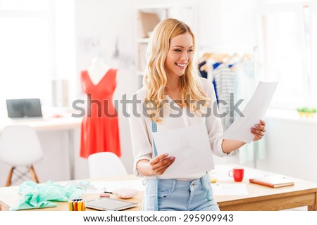 Looking over some designs. Cheerful young woman holding papers with drawings and smiling while leaning at the desk in fashion workshop