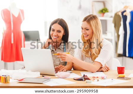 Researching the new fashion trends. Two joyful young women working together while sitting at the desk in their fashion workshop