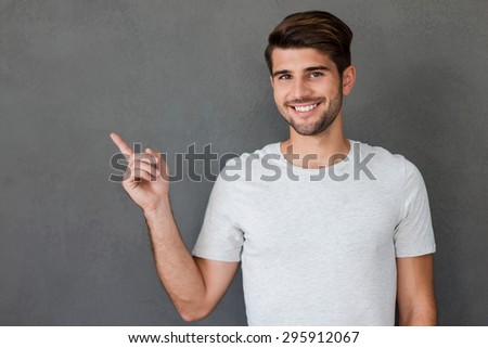 Look at that! Happy young man pointing away and smiling while standing against grey background