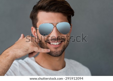 Call me! Happy young man gesturing mobile phone near face and smiling while standing against grey background