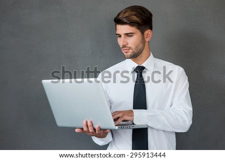 Confident business expert. Confident young man in shirt and tie working on laptop while standing against grey background