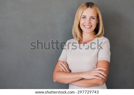 Confident beauty. Cheerful mature woman keeping arms crossed and smiling while standing against grey background