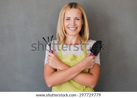 Ready for gardening. Happy mature woman in green apron holding gardening equipment and smiling while standing against grey background