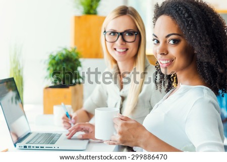 Two minds are better than one. Two smiling young women looking at camera and smiling while sitting at working place