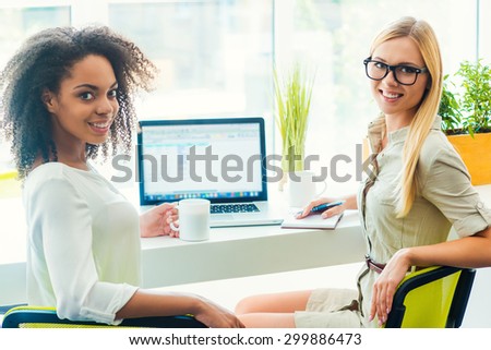 Teamwork makes great work. Two cheerful young women looking at camera and smiling while sitting at working place