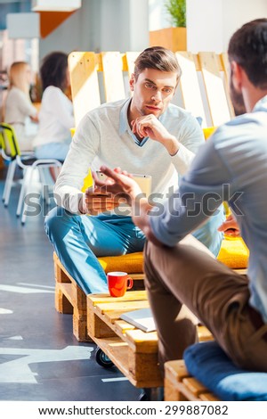 Business conversation. Two confident young men in smart casual wear discussing something while sitting in office with their colleagues working in the background