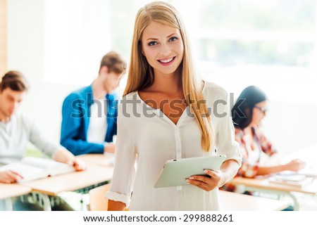 Tablet for making her study easier. Smiling young woman holding digital tablet and looking at camera while her classmates studying in the background