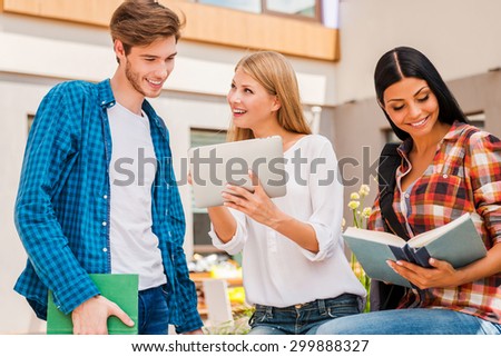 Campus life is awesome! Smiling young woman showing something on digital tablet to young man while young woman reading a book