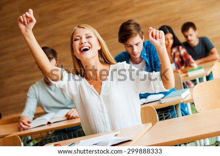 I did it! Excited young woman keeping arms raised while sitting at front desk in the classroom