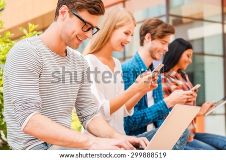 Living in digital age. Group of young people holding different digital devices and smiling while sitting in a row outdoors