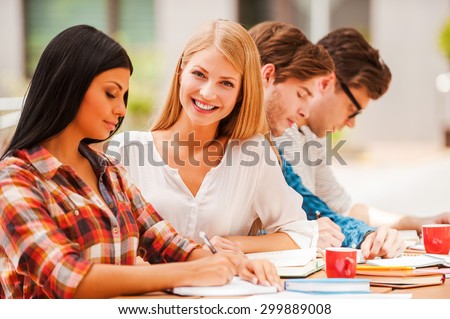 Finding inspiration in friends. Happy young woman smiling and looking at camera while sitting with her friends at the wooden desk outdoors