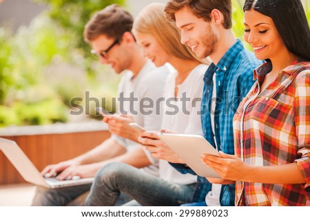 Digital world. Group of happy young people holding different digital devices and smiling while sitting in a row outdoors