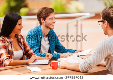 Good time with college friends. Group of cheerful young people smiling and discussing something while sitting at the wooden desk outdoors