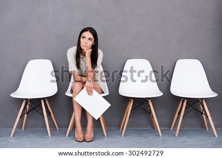 Tired of waiting. Bored young businesswoman holding paper and looking away while sitting on chair against grey background