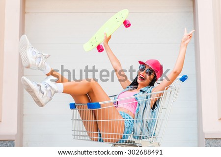 Letting go all inhibitions. Joyful young woman stretching out skateboard and gesturing peace sign while sitting in shopping cart against the garage door