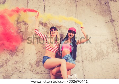 Living the bright lives. Low angle view of two cheerful young women holding smoke bombs and smiling while posing against the concrete wall
