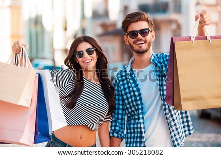 They love shopping together. Cheerful young loving couple stretching out shopping bags and smiling while walking along the street