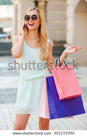 Inviting friend for shopping. Cheerful young woman holding shopping bags and talking on the phone while standing outdoors