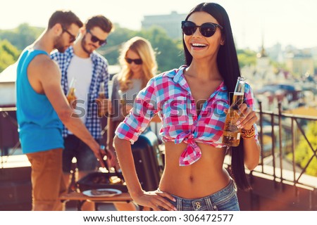 Chill time with friends. Happy young woman holding bottle with beer and smiling while three people barbecuing in the background